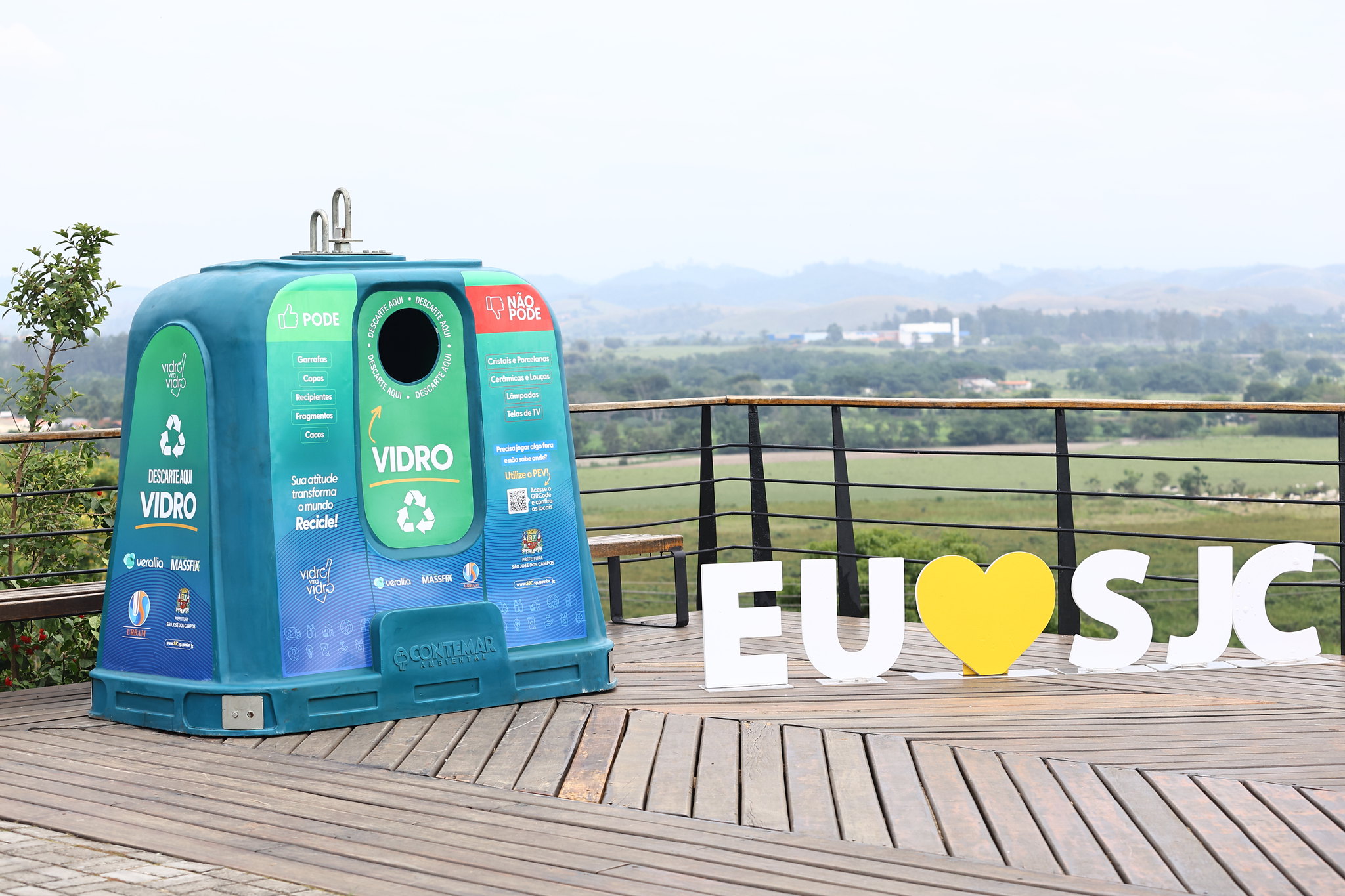 Ecoponto para descarte de vidro ao lado de placa Eu Amo SJC no deck da Anchieta com banhado ao fundo